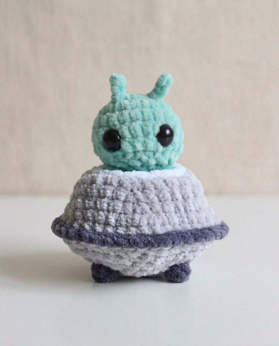 Plush crochet amigurumi alien with big black eyes sitting inside its grey and green flying saucer