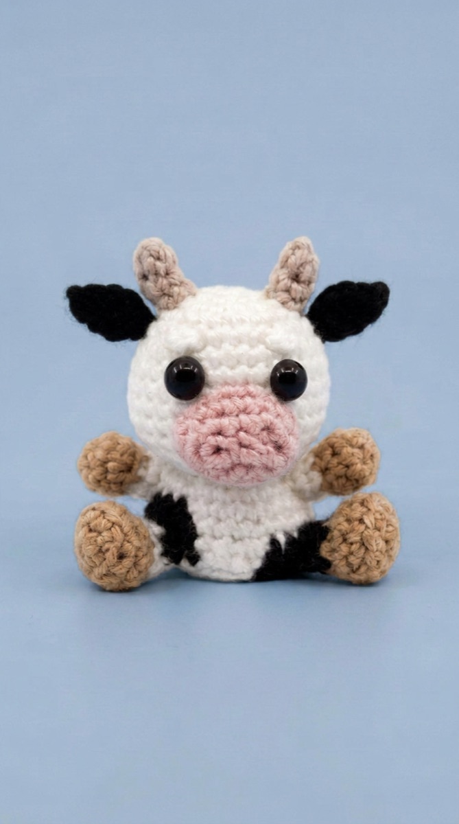 Tiny crochet cow amigurumi sitting up with black ears and tan legs