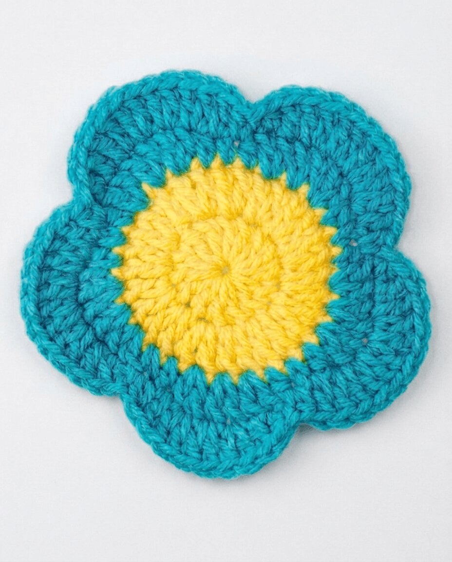 Crochet flower coaster with a yellow center and five rounded teal petals, lying flat on a white surface