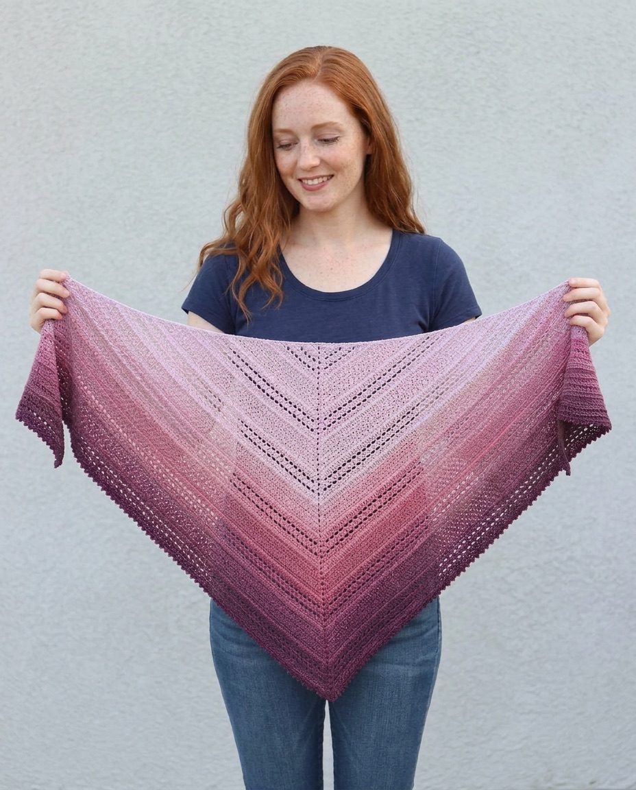 Berry pink and burgundy gradient crochet triangle shawl with ridged rows and a picot edge, held open against a wall